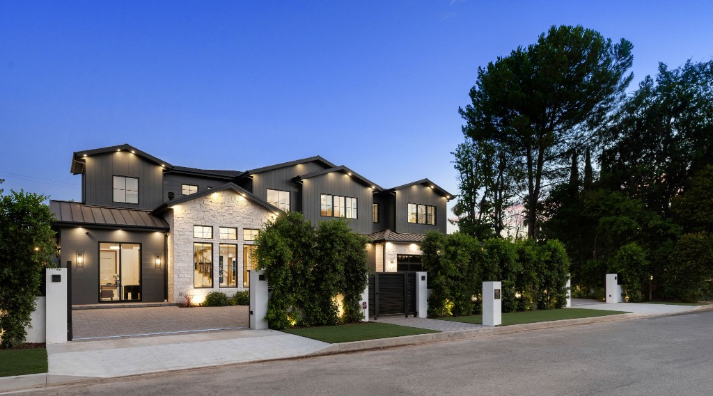 Exterior of a large modern luxury home with dark gray siding, white stone accents, and lush green landscaping, beautifully illuminated at dusk